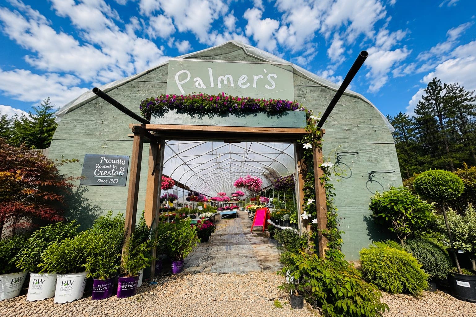 The Palmer's greenhouse entrance under a blue sky, with a hand-painted sign, flower boxes, and a 'Proudly Rooted in Creston Since 1983' plaque